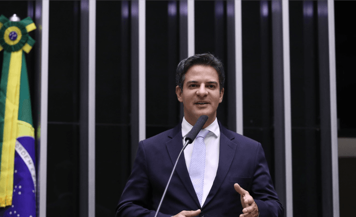 Deputado discursa no plenário da Câmara com a bandeira do Brasil ao fundo.