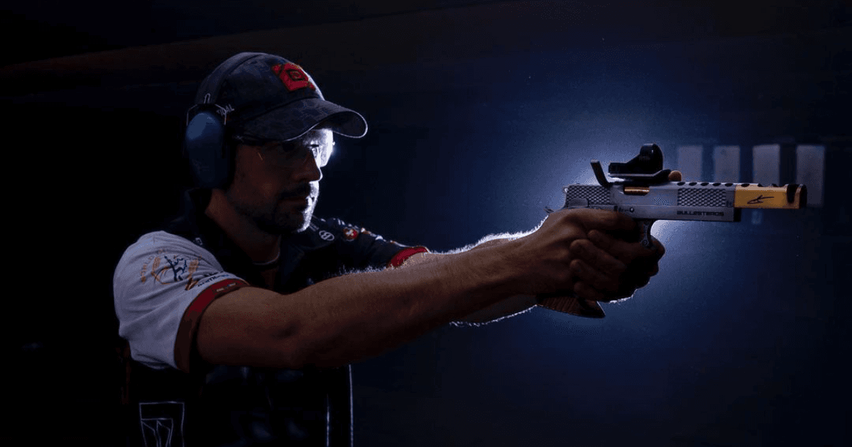 Jorge Ballesteros atira com uma pistola equipada com mira em ambiente escuro.