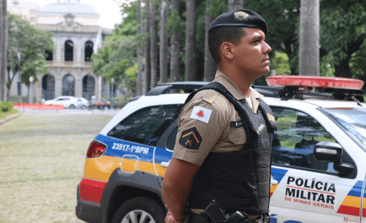 Policial militar de Minas Gerais diante de viatura e prédio público.