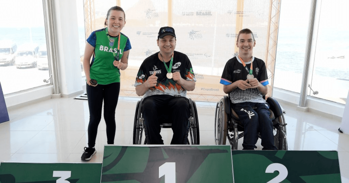 Três atletas do tiro paralímpico exibem medalhas em um pódio.