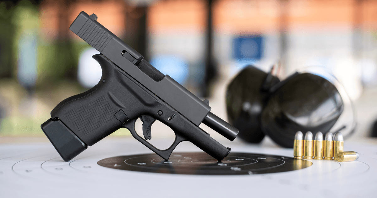 Pistola preta, óculos de proteção e cartuchos sobre um alvo de papel.