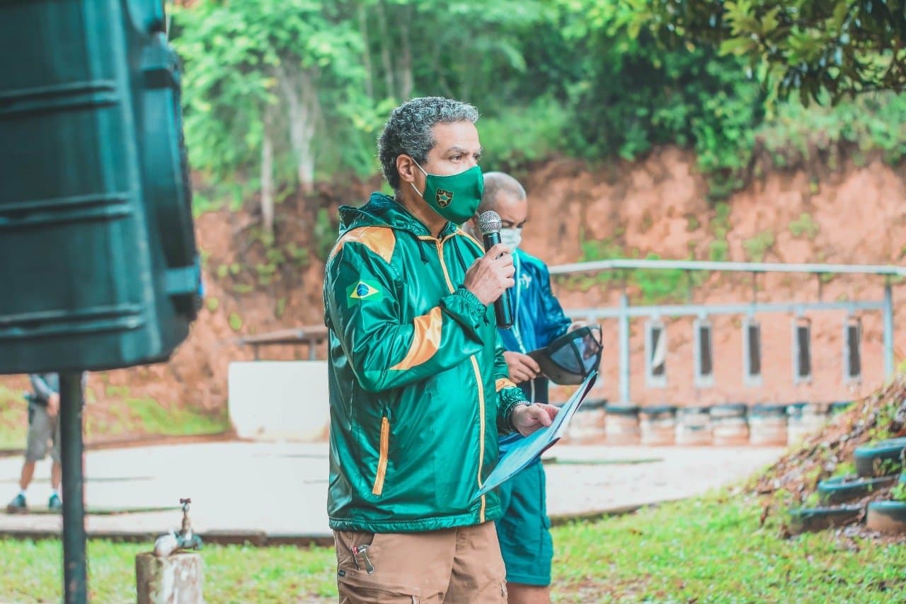 Demetrius Oliveira fala ao microfone em evento de tiro ao ar livre.