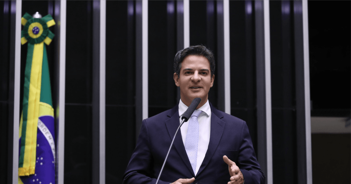 Deputado discursa no plenário da Câmara com a bandeira do Brasil ao fundo.