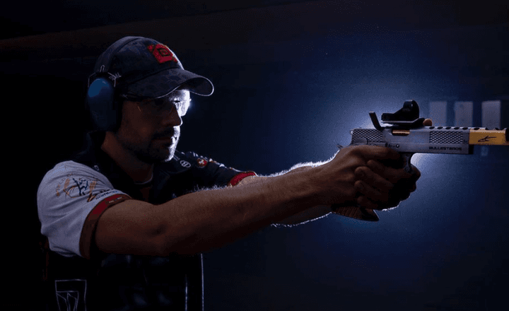 Jorge Ballesteros atira com uma pistola equipada com mira em ambiente escuro.