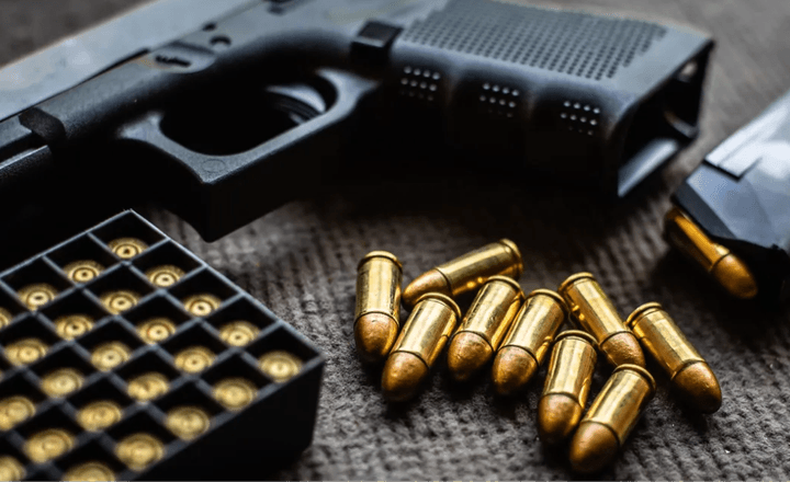 Pistola, caixa de munição e cartuchos espalhados sobre uma mesa escura.