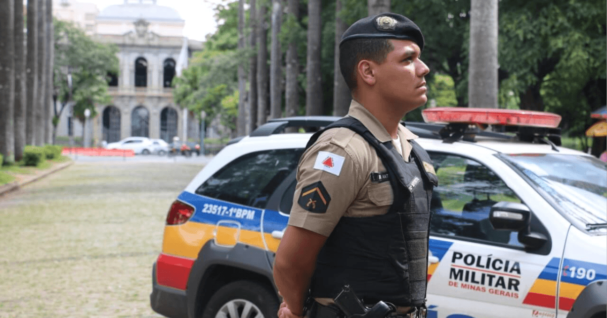 Policial militar de Minas Gerais diante de viatura e prédio público.