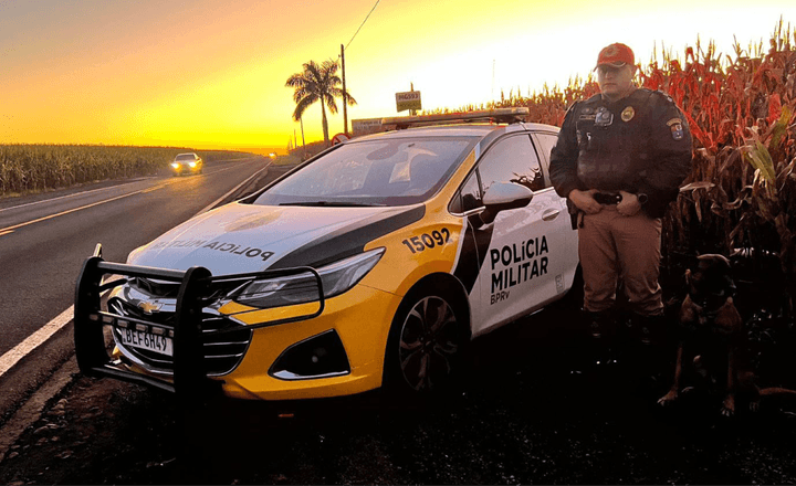 Policial militar ao lado de viatura em rodovia durante o pôr do sol.