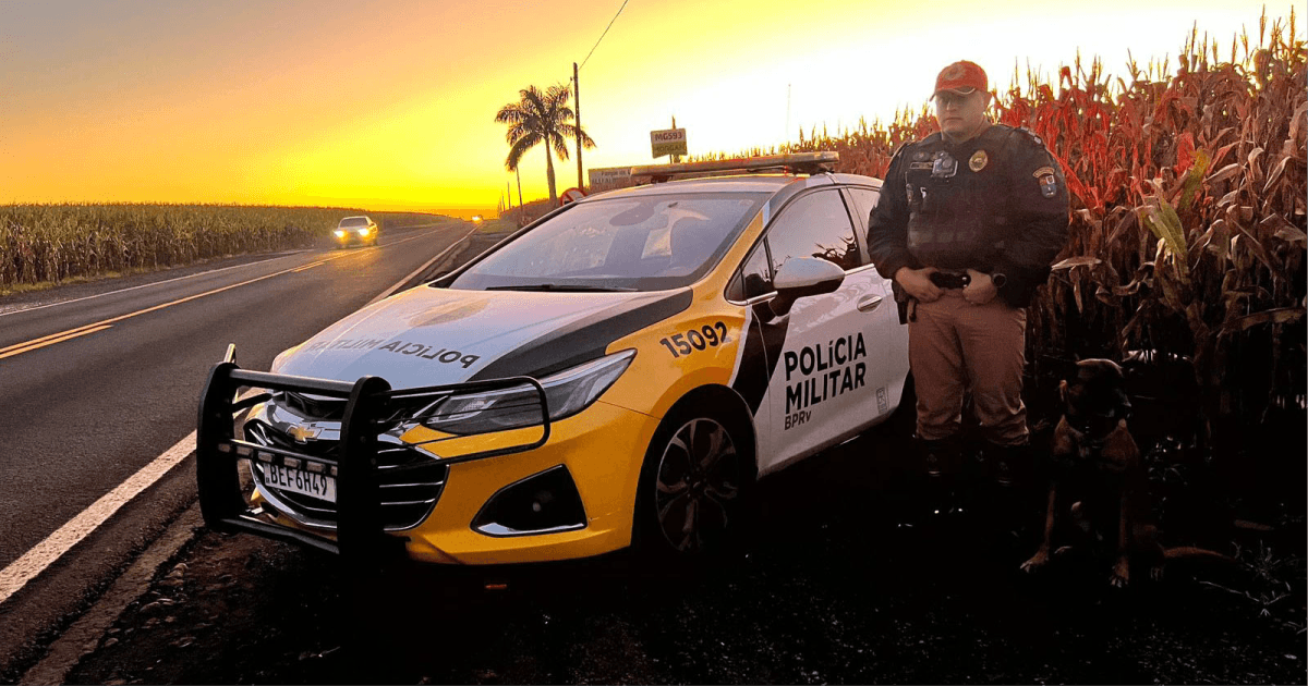 Policial militar ao lado de viatura em rodovia durante o pôr do sol.