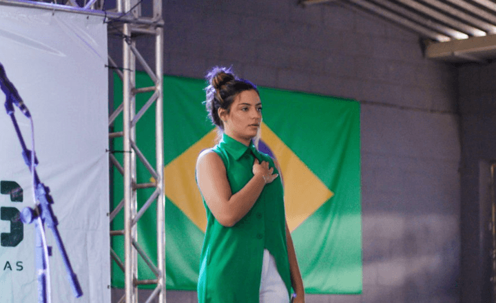 Mulher fala ao microfone em palco diante de uma bandeira do Brasil.
