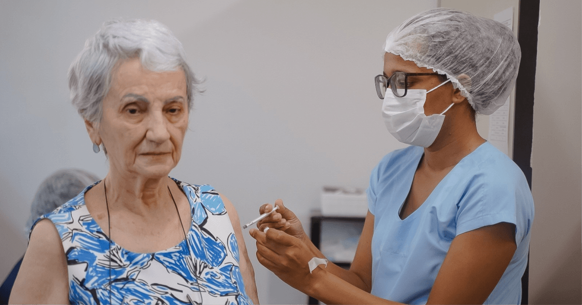 Profissional de saúde aplica vacina no braço de uma mulher idosa.
