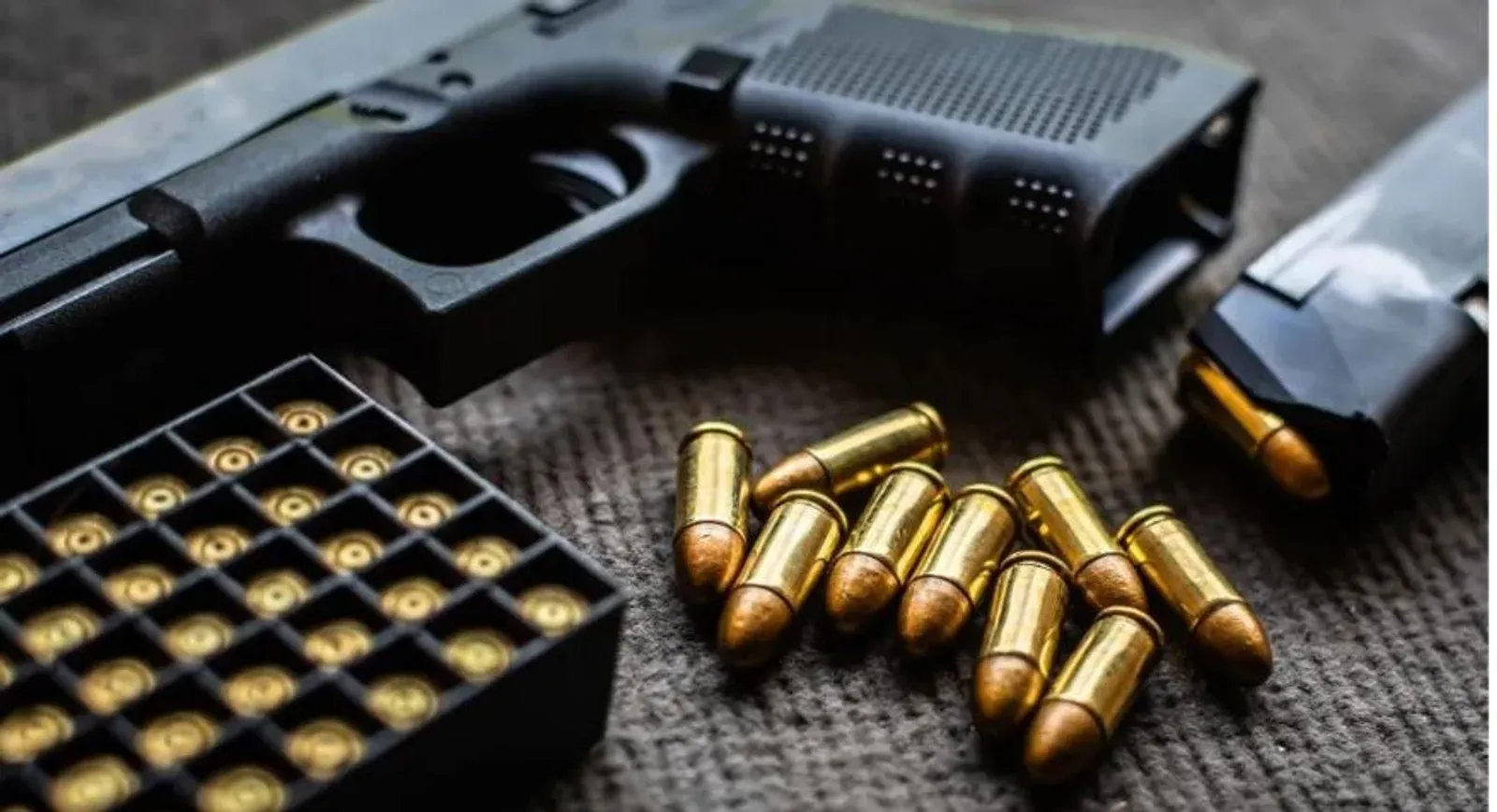 Pistola, caixa de munição e cartuchos espalhados sobre uma mesa escura.